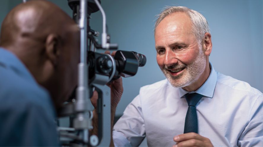 A-man-looking-at-a-camera-while-another-man-undergoes-an-eye-exam