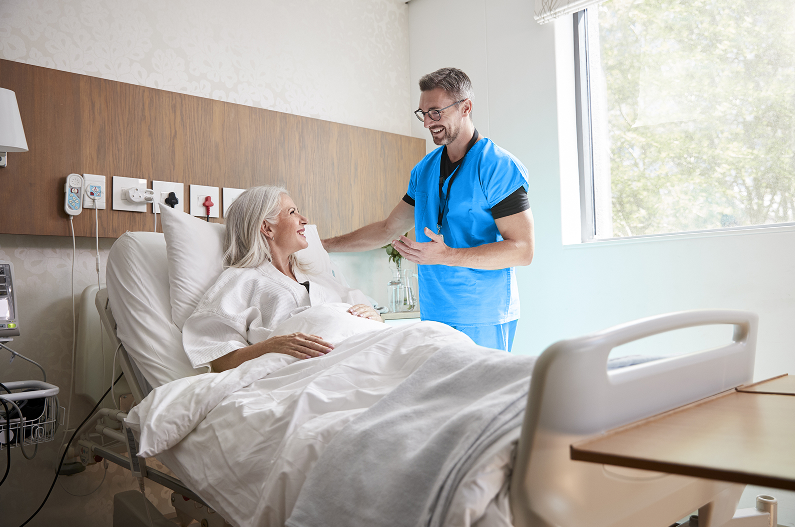 Senior female patient in hospital bed talking to male in blue scrubs