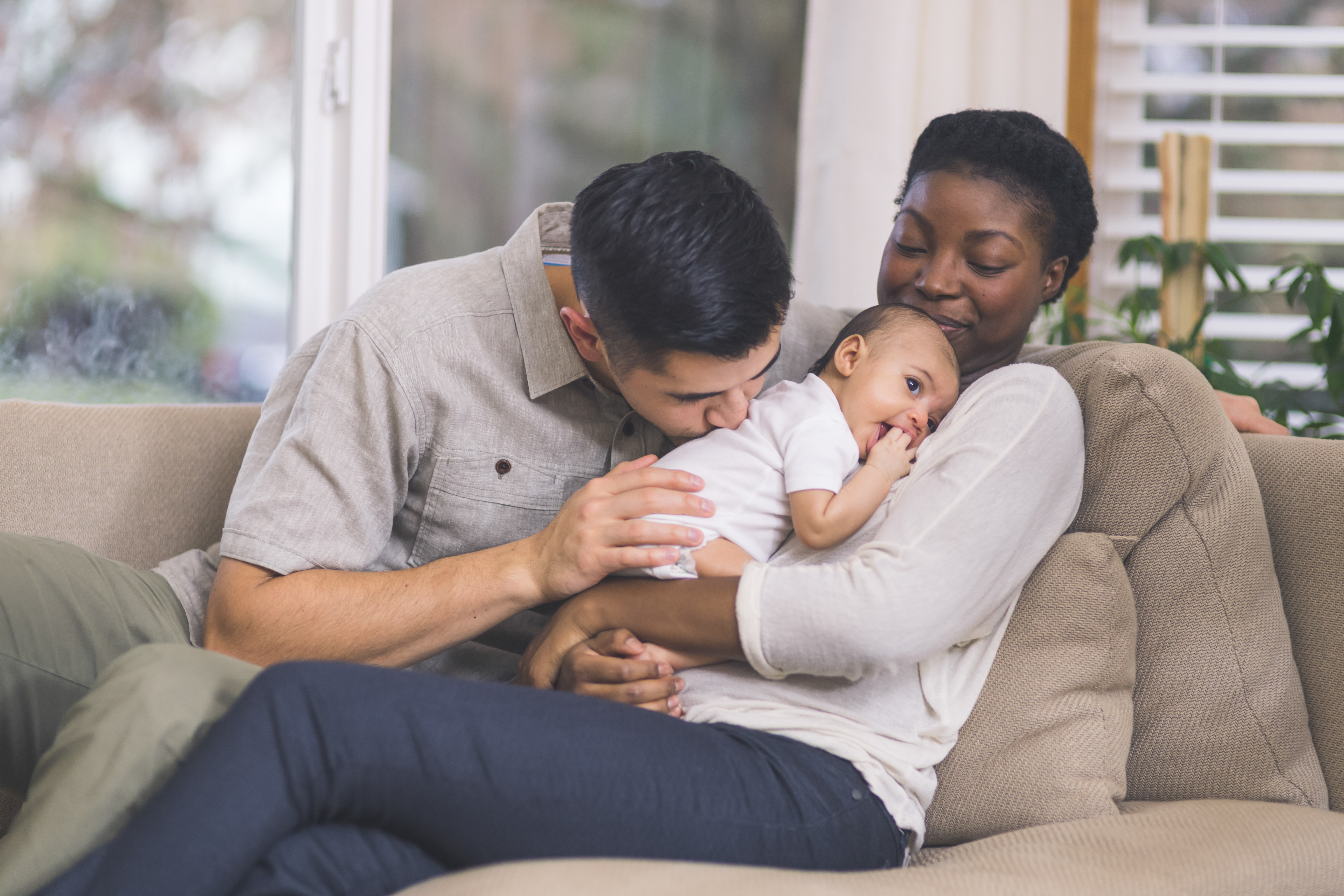 Interracial couple snuggle with their infant son on the couch in their living room
